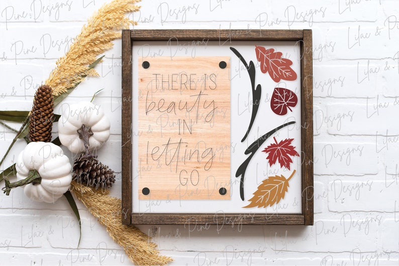 May include: A wooden sign with a brown frame and a white background. The sign has a quote that reads "There is beauty in letting go." The quote is written in black script font. The sign is decorated with fall leaves in shades of red, orange, and yellow.