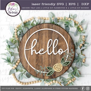 May include: A wooden round sign with the word "hello" in white script inside a white circle. The sign is decorated with a green floral wreath with a yellow rose.