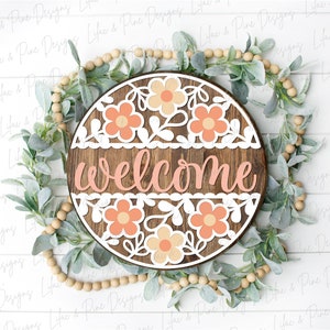 May include: A round wooden sign with the word "welcome" in pink lettering. The sign is decorated with white and peach paper flowers and leaves.