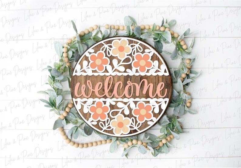 May include: A round wooden sign with the word "welcome" in pink lettering. The sign is decorated with white and peach paper flowers and leaves.