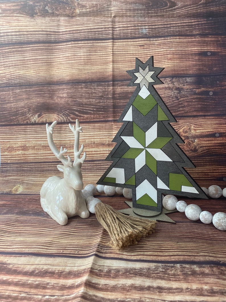 Op de afbeelding: Een houten kerstboomdecoratie met een geometrisch patroon in groen, wit en zwart. De boom staat op een houten oppervlak met een wit keramiek rendierbeeldje en een bruine kwast.