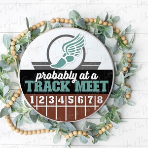 May include: A round sign with the text "probably at a TRACK MEET" in teal and white. The sign features a track shoe with wings and a track with numbers 1-8. The sign is surrounded by greenery and wooden beads.