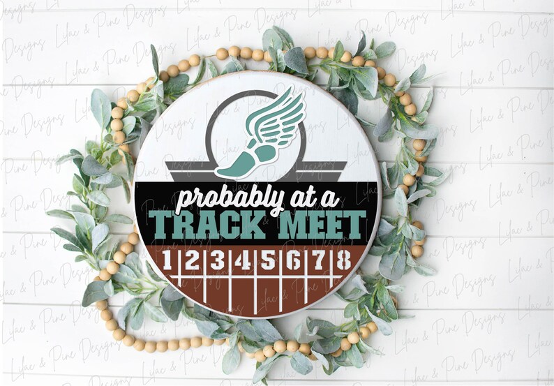 May include: A round sign with the text "probably at a TRACK MEET" in teal and white. The sign features a track shoe with wings and a track with numbers 1-8. The sign is surrounded by greenery and wooden beads.