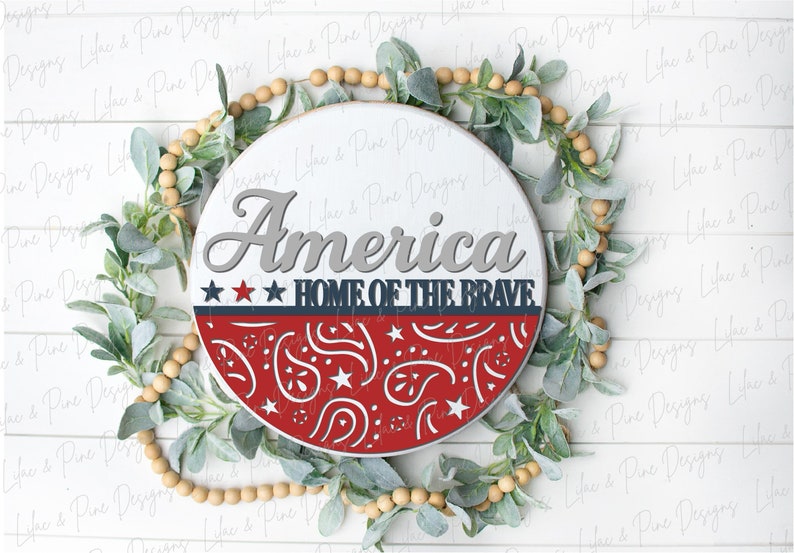 Puede incluir: Un letrero redondo de madera con las palabras "America Home of the Brave" en letras grises. El letrero est&aacute; decorado con un patr&oacute;n de bandana rojo, blanco y azul.