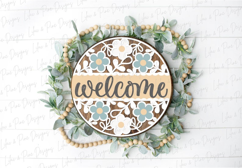 May include: A round wooden sign with a welcome message in gray lettering. The sign is decorated with a white lace border and blue and white flowers.