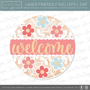 May include: A round wooden sign with a pink welcome banner and white floral cutouts. The sign is decorated with red, pink, and blue flowers.
