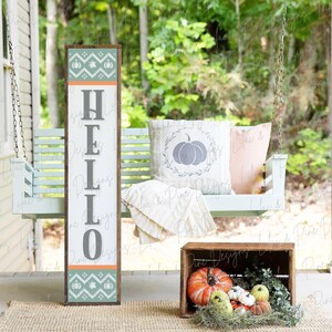 May include: A wooden sign with the word "HELLO" in gray letters on a white background. The sign has a teal and orange patterned border with pumpkins. The sign is leaning against a white porch swing with a pillow that has a gray pumpkin design. A wooden crate with orange pumpkins and greenery is on the porch floor.