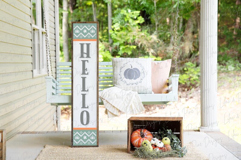 May include: A wooden sign with the word "HELLO" in gray letters on a white background. The sign has a teal and orange patterned border with pumpkins. The sign is leaning against a white porch swing with a pillow that has a gray pumpkin design. A wooden crate with orange pumpkins and greenery is on the porch floor.