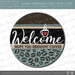 K&ouml;nnte beinhalten: Ein rundes Holzschild mit einem Kaffeetassen- und Dampfdesign, einem schwarzen Hintergrund und dem Text "Welcome Hope You Brought Coffee". Das Schild ist mit einem braunen und blauen Kaffeekornmuster verziert.