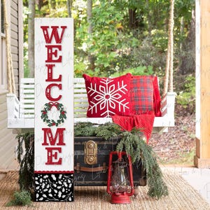 May include: A white wooden sign with the word "WELCOME" in red letters. The sign has a wreath with red berries and green leaves in the center. The sign is decorated with a black and white pattern at the bottom.