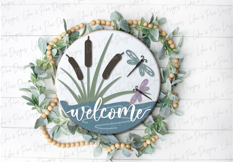 May include: A round wooden sign with a blue and white water scene, green cattails, and two purple and blue dragonflies. The sign says "welcome" in white cursive lettering.