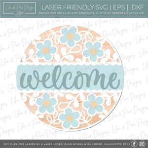 May include: A round wooden sign with a laser-cut design of blue flowers and leaves. The sign says "welcome" in a light blue font.