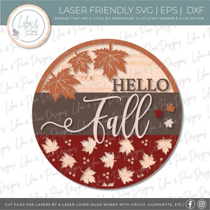 May include: A round wooden sign with a brown, gray, and red background. The sign features the text "Hello Fall" in white script with a brown outline. The sign is decorated with fall leaves in shades of orange, red, and brown.