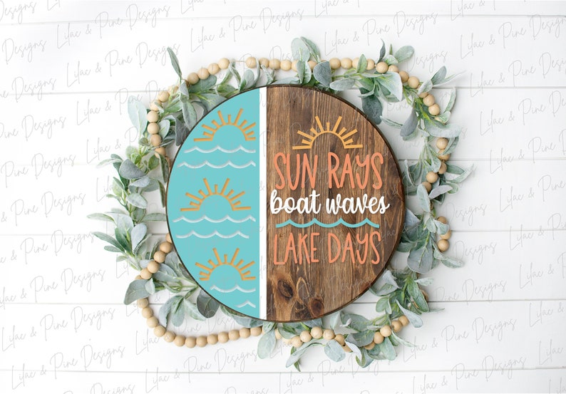 Op de afbeelding: Een rond houten bord met een blauwe en bruine achtergrond. De linkerzijde van het bord heeft een turquoise blauwe achtergrond met drie gele zonnen en witte golven. De rechterzijde van het bord heeft een bruine houten achtergrond met de tekst "SUN RAYS boat waves LAKE DAYS" in oranje en witte letters.