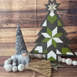 Op de afbeelding: Een geometrische, witte, groene en bruine houten kerstboomversiering met een witte ster bovenop. Een grijze gnome met een zwart-witte gepatenteerde hoed en een witte baard zit voor de boom. De gnome houdt een bruin en beige bezem vast. De boom en de gnome staan op een houten oppervlak met witte kralen.