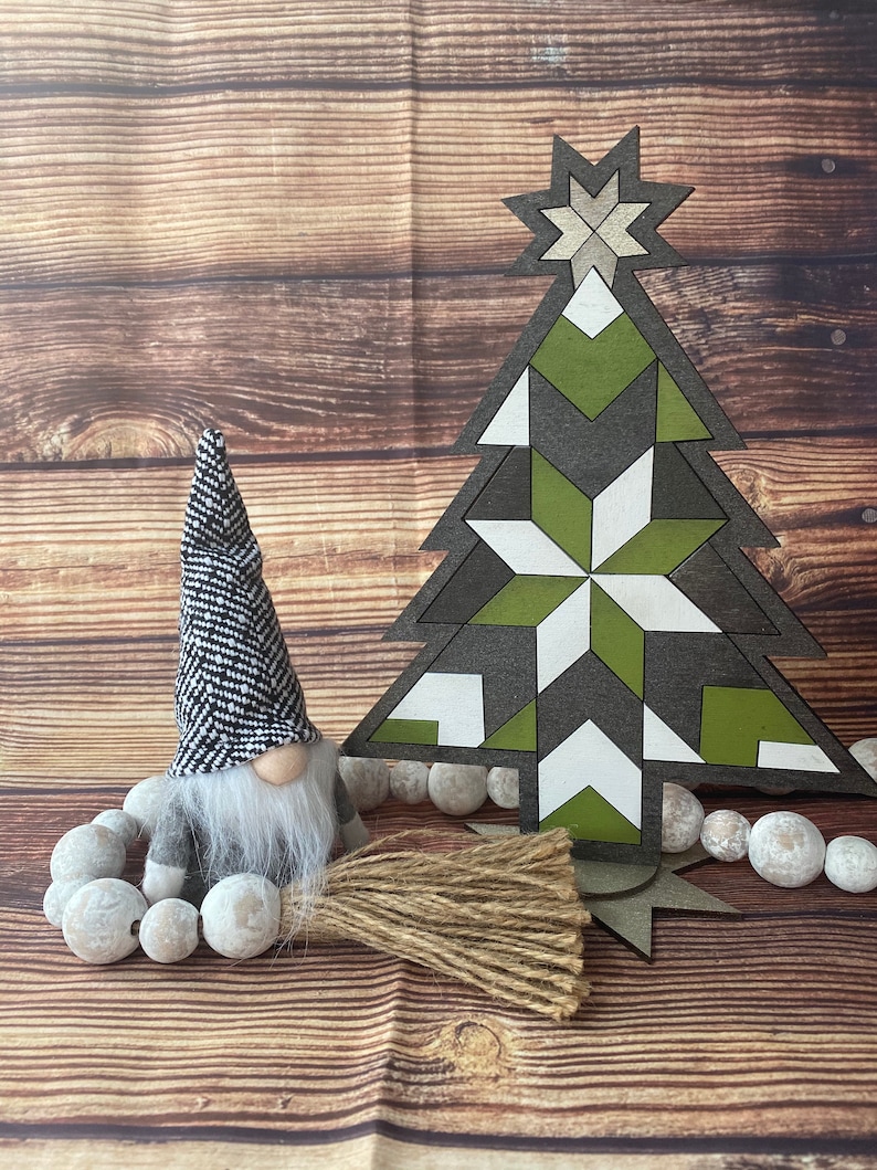 Op de afbeelding: Een geometrische, witte, groene en bruine houten kerstboomversiering met een witte ster bovenop. Een grijze gnome met een zwart-witte gepatenteerde hoed en een witte baard zit voor de boom. De gnome houdt een bruin en beige bezem vast. De boom en de gnome staan op een houten oppervlak met witte kralen.
