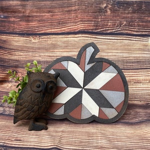May include: A wooden pumpkin shaped ornament with a geometric pattern in shades of brown, gray, white, and red. The ornament is sitting on a wooden surface with a small cast iron owl figurine next to it.