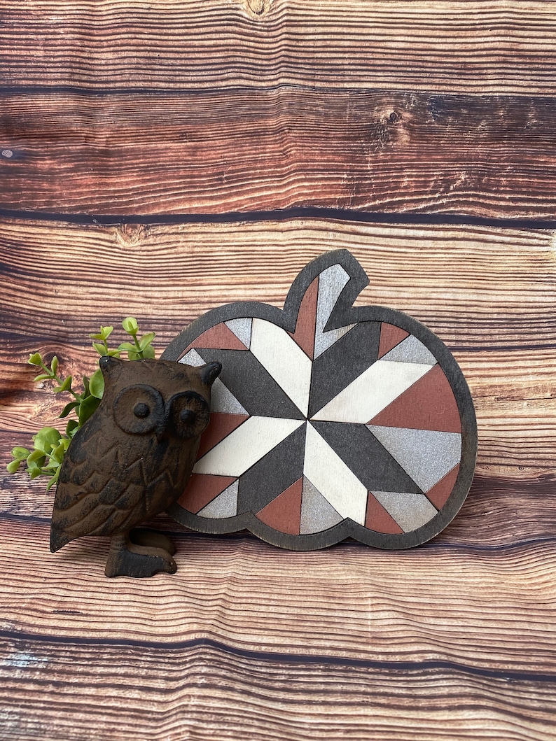 May include: A wooden pumpkin shaped ornament with a geometric pattern in shades of brown, gray, white, and red. The ornament is sitting on a wooden surface with a small cast iron owl figurine next to it.