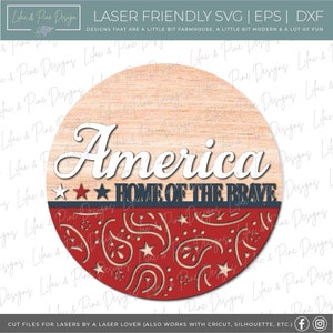 Puede incluir: Un letrero redondo de madera con un dise&ntilde;o de bandera estadounidense roja, blanca y azul. El letrero dice "America Home of the Brave" en letras blancas.