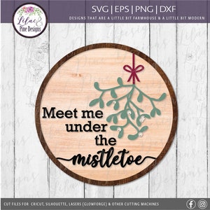 May include: A round wooden sign with a light wood grain background and a dark brown border. The sign features the text "Meet me under the mistletoe" in black, with a sprig of green mistletoe and a red bow. The sign is designed for cutting machines.