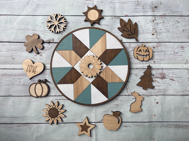 K&ouml;nnte beinhalten: Ein rundes Holzschild mit einem geometrischen Muster in Blau, Wei&szlig; und Braun. Das Schild ist von verschiedenen Holz-Ausschnitten umgeben, darunter eine Sonnenblume, ein Stern, ein K&uuml;rbis, ein Blatt, eine Schneeflocke, ein Herz mit dem Wort "LOVE" darauf, ein Kleeblatt, eine Sonne, ein K&uuml;rbis mit einem Gesicht, ein Weihnachtsbaum mit dem Wort "JOY" darauf, ein Kaninchen und ein Apfel.