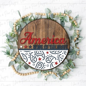Puede incluir: Un letrero redondo de madera con las palabras "America Home of the Brave" en letras rojas y azules. El letrero est&aacute; decorado con un patr&oacute;n paisley blanco y azul.