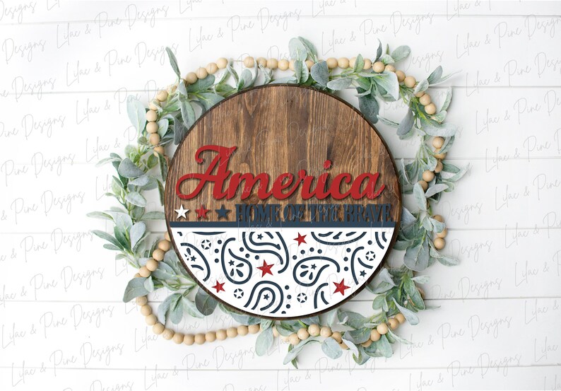 Puede incluir: Un letrero redondo de madera con las palabras "America Home of the Brave" en letras rojas y azules. El letrero est&aacute; decorado con un patr&oacute;n paisley blanco y azul.