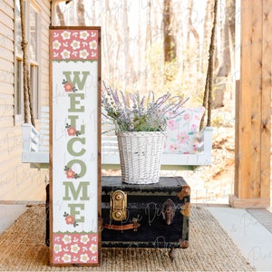 May include: A wooden welcome sign with a green welcome message and pink and white floral accents. The sign is leaning against a black vintage trunk on a porch with a white swing and a potted plant.