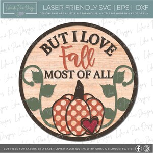 May include: A round wooden sign with the text "BUT I LOVE FALL MOST OF ALL" in brown and orange. The sign features a pumpkin with polka dots and a heart, along with green leaves. The design is suitable for laser cutting.