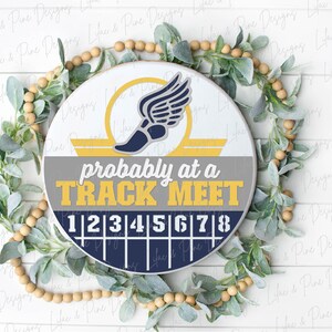 May include: A round wooden sign with the text "probably at a TRACK MEET" in yellow and gray. The sign features a track shoe with wings and a track lane design. The sign is surrounded by a green and beige wreath.