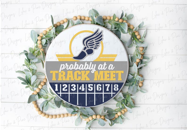 May include: A round wooden sign with the text "probably at a TRACK MEET" in yellow and gray. The sign features a track shoe with wings and a track lane design. The sign is surrounded by a green and beige wreath.