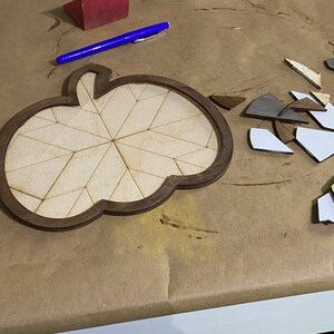 May include: A wooden pumpkin-shaped puzzle with a brown frame and a light brown interior. The puzzle is made up of many small pieces that are scattered around the frame.