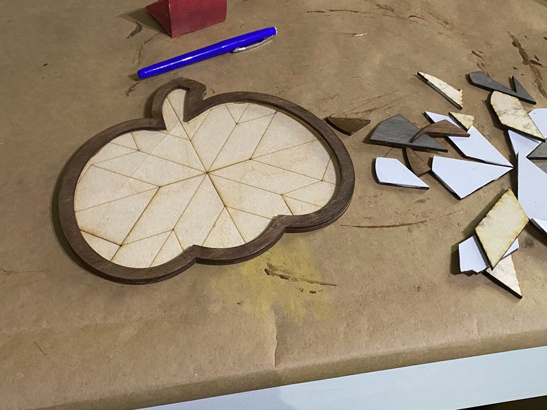 May include: A wooden pumpkin-shaped puzzle with a brown frame and a light brown interior. The puzzle is made up of many small pieces that are scattered around the frame.