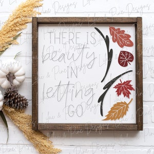 May include: A wooden sign with a white background and brown frame. The sign has black calligraphy that reads "There is beauty in letting go."  The calligraphy is surrounded by a black vine with red, orange, and brown leaves.