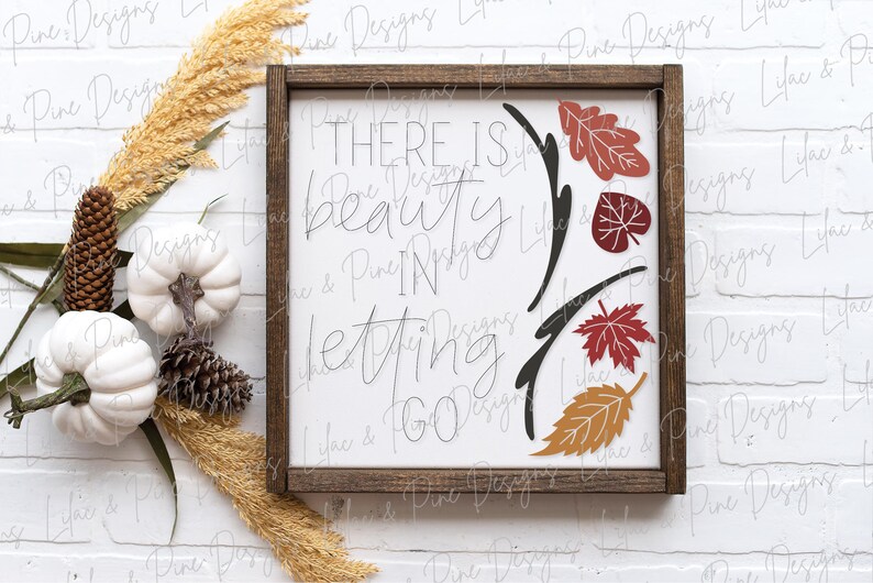 May include: A wooden sign with a white background and brown frame. The sign has black calligraphy that reads "There is beauty in letting go."  The calligraphy is surrounded by a black vine with red, orange, and brown leaves.