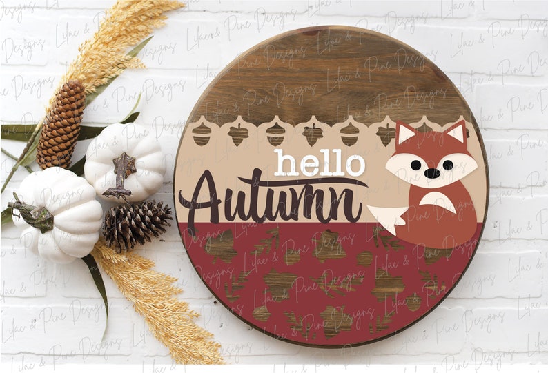 May include: A round wooden sign with a brown and tan background. The sign says "hello autumn" in white letters with a brown fox sitting on the bottom right corner. The sign is decorated with acorns and leaves.