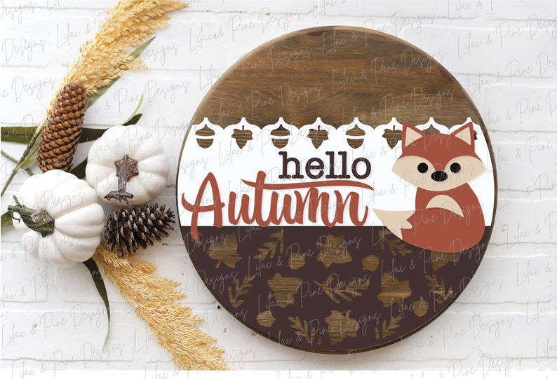 May include: A round wooden sign with a brown and white background. The sign says "hello autumn" in red and white lettering. A brown fox with white accents is on the right side of the sign.