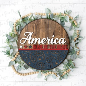 Puede incluir: Un letrero redondo de madera con las palabras "America Home of the Brave" en letras blancas. El letrero est&aacute; decorado con estrellas rojas, blancas y azules y un dise&ntilde;o azul y blanco.