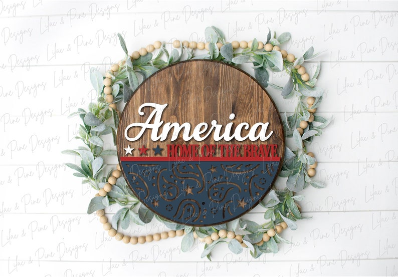 Puede incluir: Un letrero redondo de madera con las palabras "America Home of the Brave" en letras blancas. El letrero est&aacute; decorado con estrellas rojas, blancas y azules y un dise&ntilde;o azul y blanco.