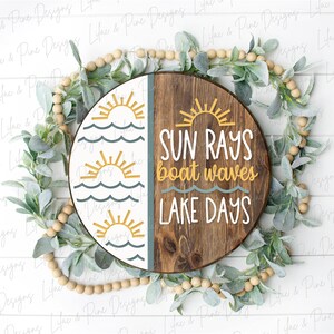 Op de afbeelding: Een rond houten bord met een witte en blauwe helft met gestileerde zonnen en golven. De andere helft is van bruin hout met de tekst "SUN RAYS boat waves LAKE DAYS" in wit.