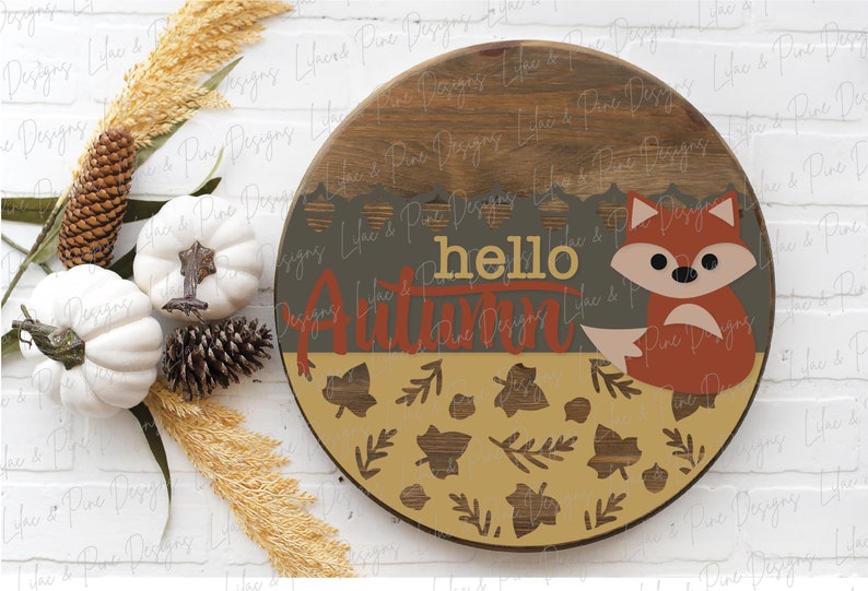 May include: A round wooden sign with a brown woodgrain finish. The sign features a painted fox with a gray face and orange body, surrounded by leaves and the text "hello autumn".