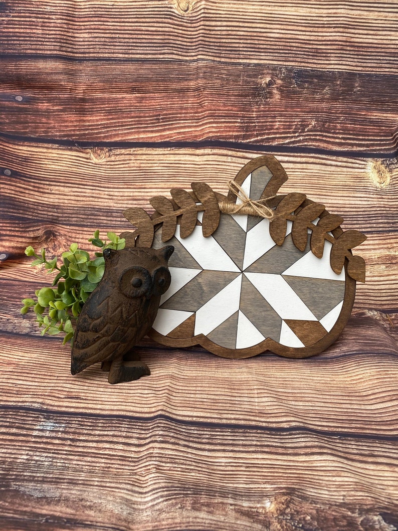 May include: A wooden pumpkin-shaped sign with a white and gray geometric pattern and a brown leafy design around the top. A small brown owl figurine is in the foreground.