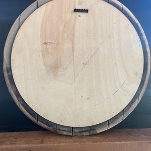 May include: Back of a round wooden barrel head with a light-colored wood center and a dark, weathered wood rim. A metal sawtooth hanger is attached to the top center. The barrel head is displayed against a dark background.