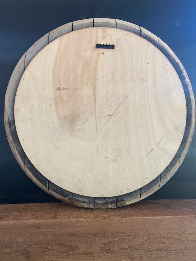 May include: Back of a round wooden barrel head with a light-colored wood center and a dark, weathered wood rim. A metal sawtooth hanger is attached to the top center. The barrel head is displayed against a dark background.