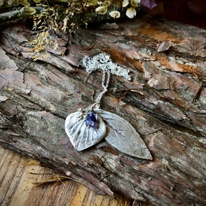 May include: A silver necklace with a leaf pendant featuring a purple gemstone. The pendant is textured and has a detailed design. The necklace is displayed on a wooden surface.