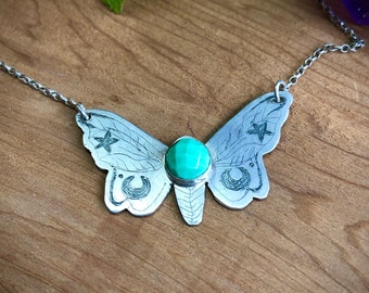 Op de afbeelding: Een zilveren ketting met een gedetailleerde mot-hanger met een turquoise edelsteen en gegraveerde sterren en halve manen.