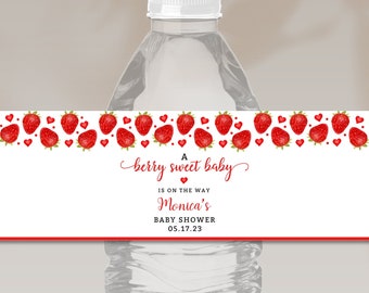 May include: A water bottle with a custom label featuring a strawberry design. The label reads "A berry sweet baby is on the way" and includes the name "Monica's" and the date "05.17.23" for a baby shower. The top of the label has a border of red strawberries.