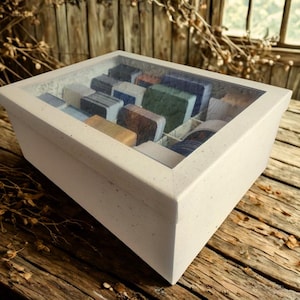 Könnte beinhalten: Eine weiße, rechteckige Box mit einem durchsichtigen Glasdeckel, gefüllt mit farbigen, rechteckigen Objekten. Die Box steht auf einer Holzoberfläche, im Hintergrund befinden sich Holzbretter und ein Fenster.