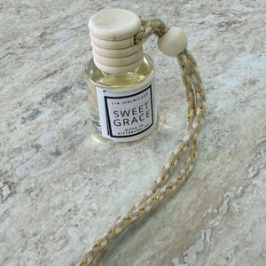 May include: A small glass bottle with a wooden cap and a braided cord. The bottle contains a light-colored liquid and a label that reads "Sweet Grace." The cord is beige with gold accents. The product is made in Pittsburgh, PA.