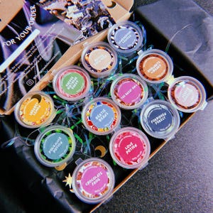 May include: A collection of 10 small round containers filled with colorful wax melts. The containers are labeled with names like "Bertie Beans", "Forbidden Forest", "Polyjuice Potion", and "Love Potion".